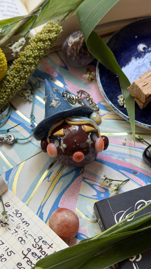 A small decorative witch figurine with a dark blue pointed hat, silver crystals and a brown body sits on a colorful patterned surface surrounded by leaves, crystals, a blue dish, and handwritten notes.