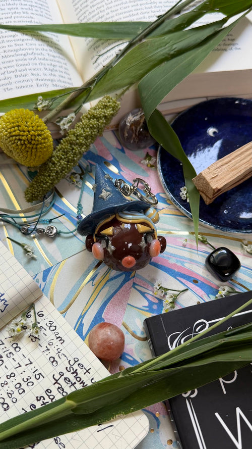 A whimsical flat-lay featuring a small witch figurine with a blue hat and silver star details, surrounded by natural elements like green leaves, a yellow flower, crystals, an open book, and a notebook on a colorful patterned surface.