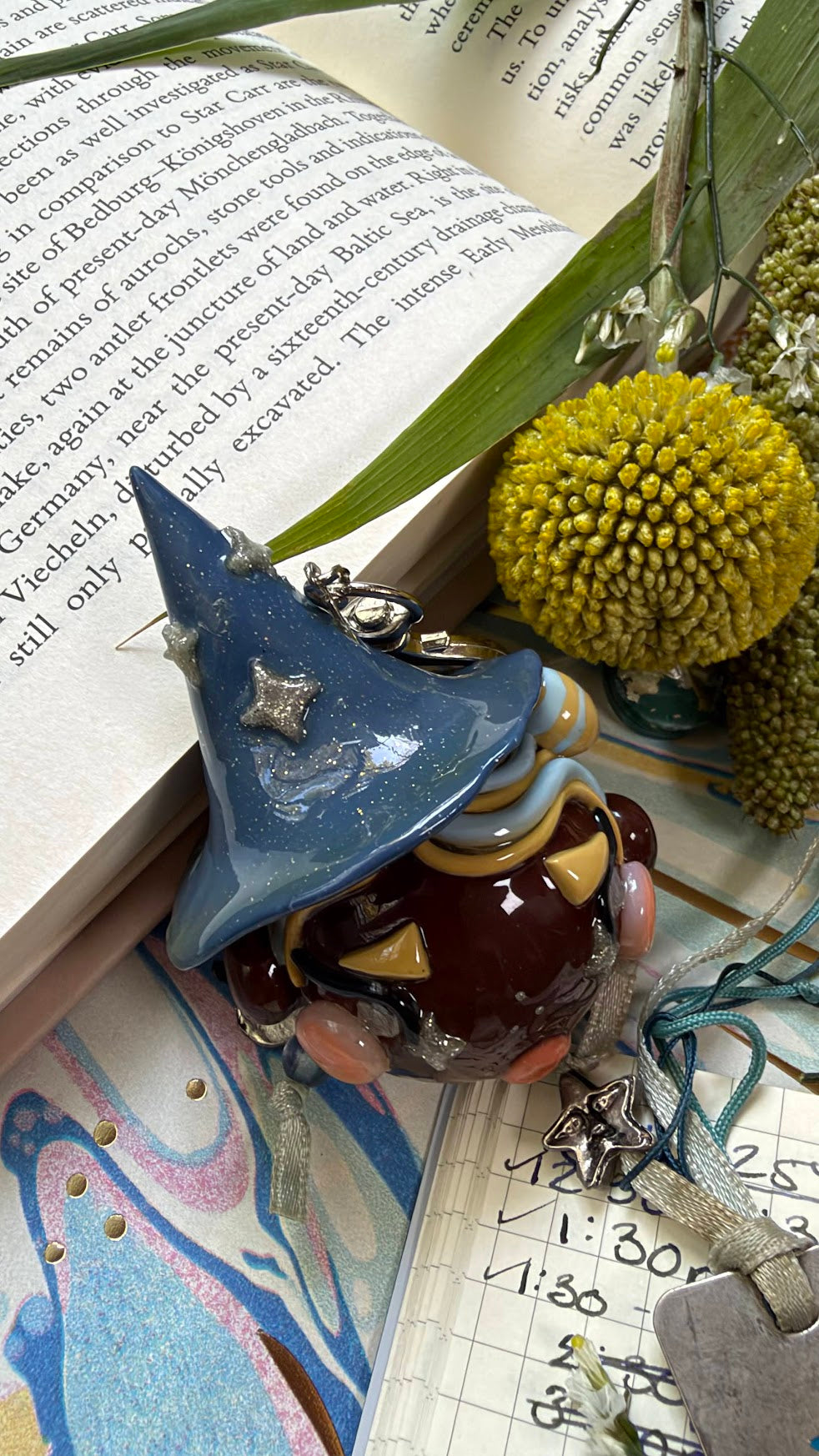 A small decorative witch figurine with a blue pointed hat and brown body, styled on top of an open book with green leaves, yellow flowers, and a notebook in the background.