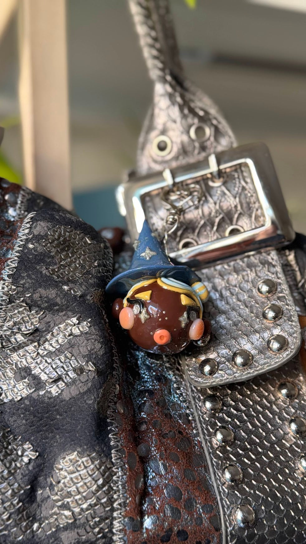 A close-up of a textured black and silver studded leather bag with a decorative brown and blue witch-themed bag charm hanging near the buckle.
