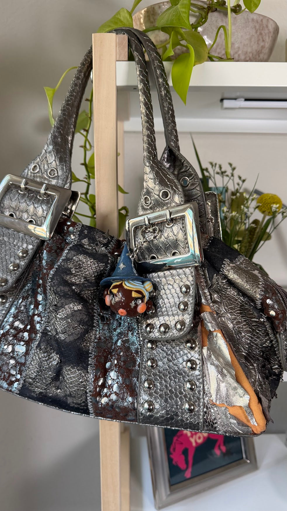 A metallic silver textured handbag with silver buckle details, studded embellishments, and a small witch with hat bag charm, hanging on a wooden stand in an indoor setting.