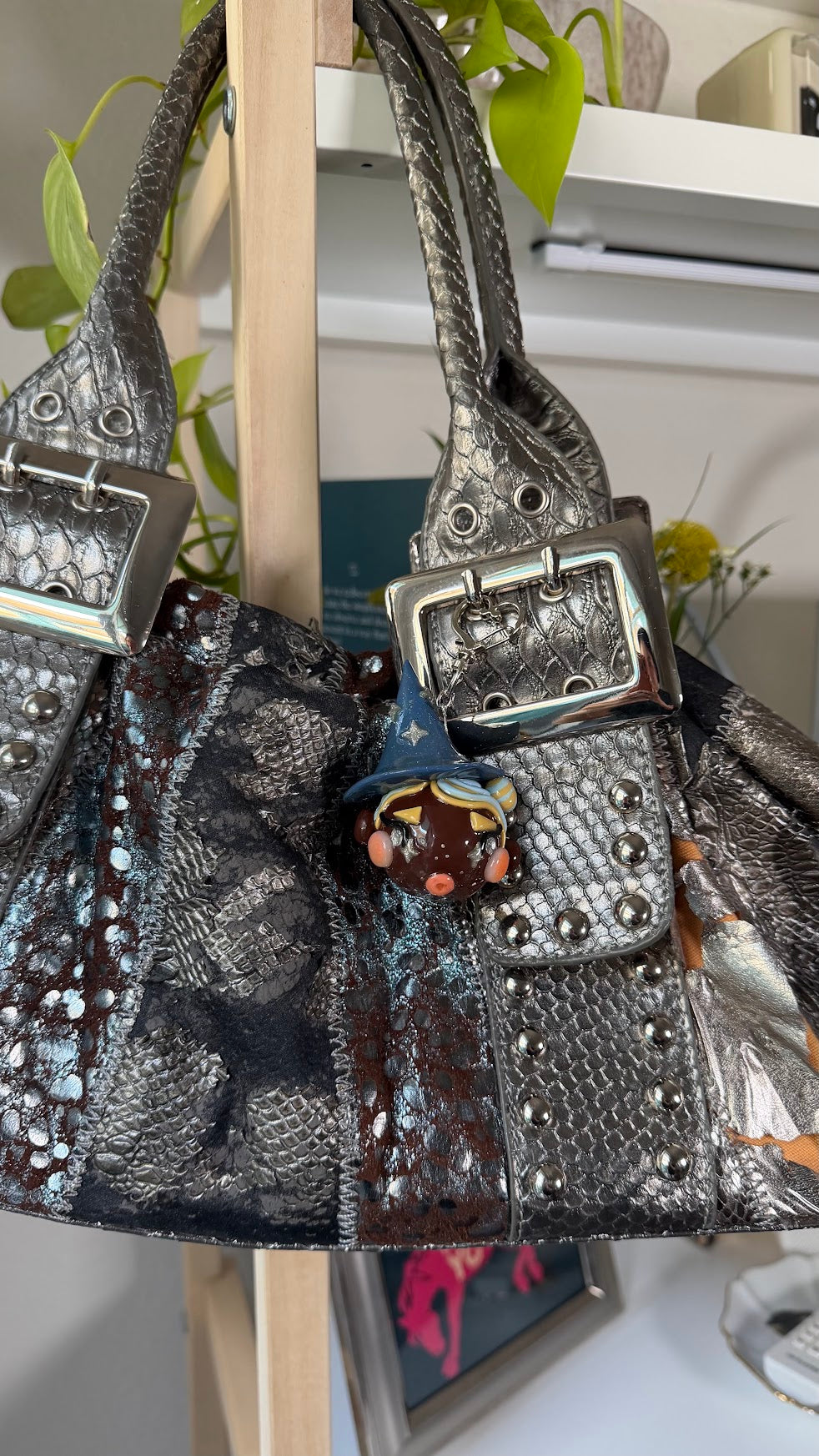 A metallic silver textured handbag with a snakeskin pattern, silver buckle details, and a small witch bag charm, hanging on a wooden display stand.