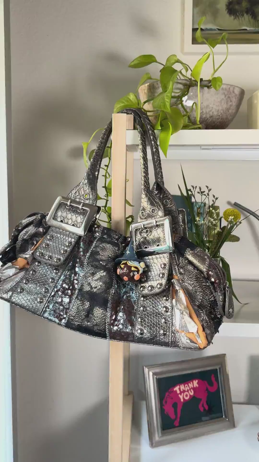 A metallic silver textured handbag with silver buckle details, studded embellishments, and a small witch with hat bag charm, hanging on a wooden stand in an indoor setting.